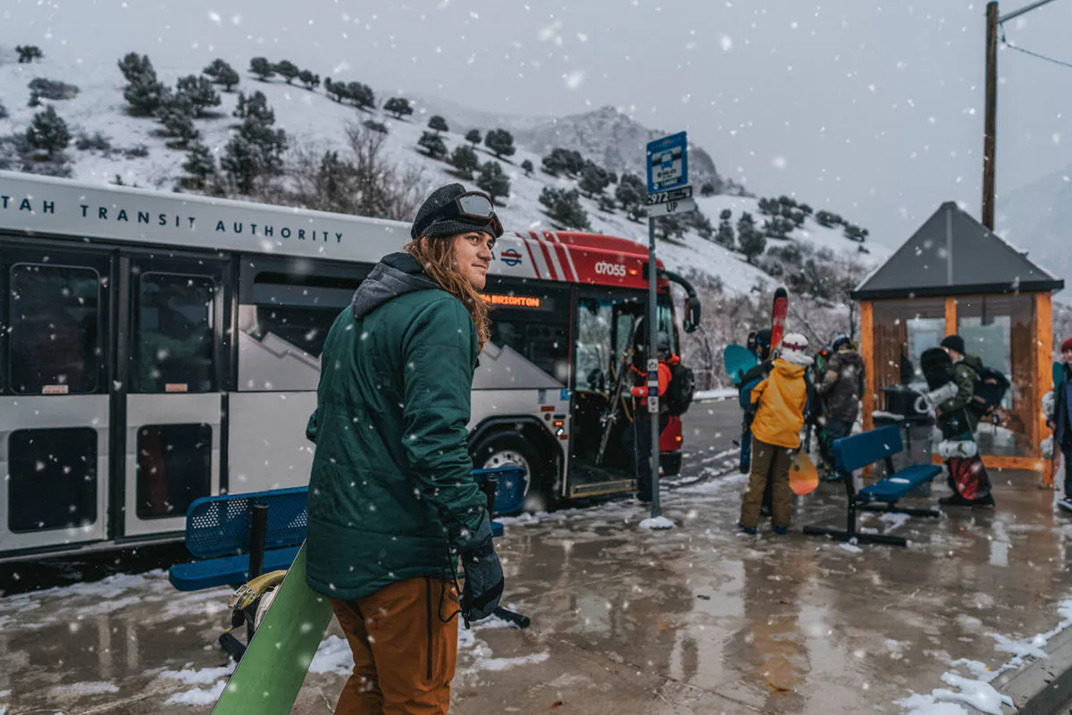 Snowy bus stop with snowboarders and skiers waiting to board the parked UTA bus. One snowboarder in green looks beyond the camera. Using a UTA ski bus will help prevent canyon congestion in the winter.