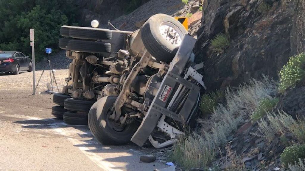An overturned dump truck rests on its side against a rock wall along SR-210 in Little Cottonwood Canyon after a speed-related crash.