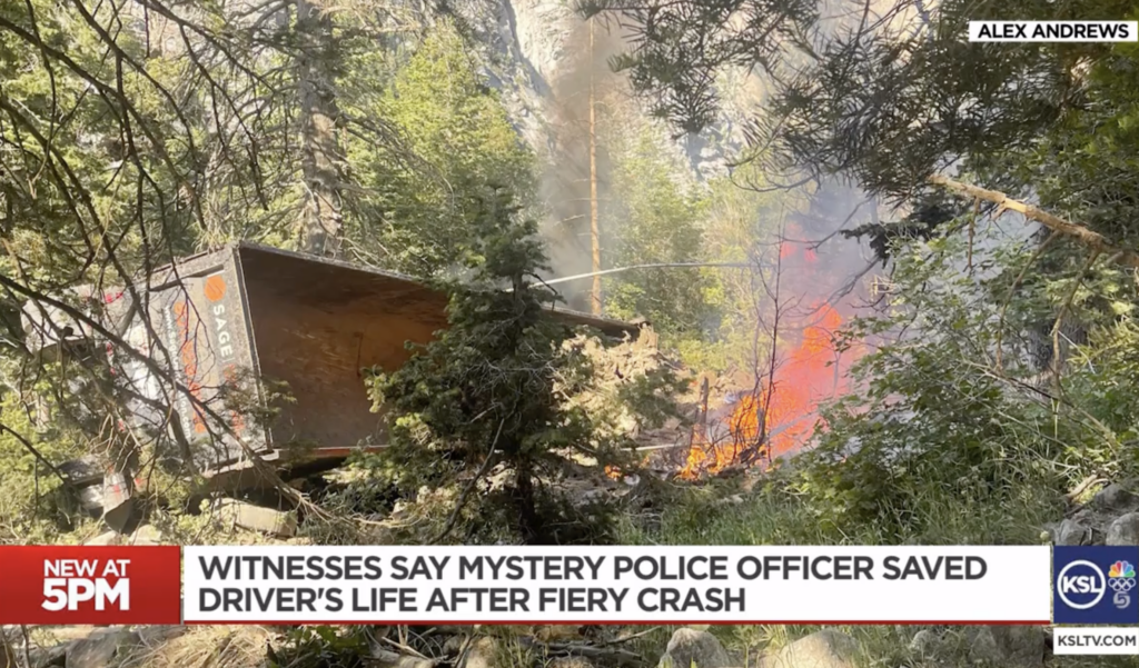 An overturned dump truck rests along SR-210 in Little Cottonwood Canyon after a speed-related crash. Flames burn through nearby vegetation and smoke rises through the surrounding trees.
