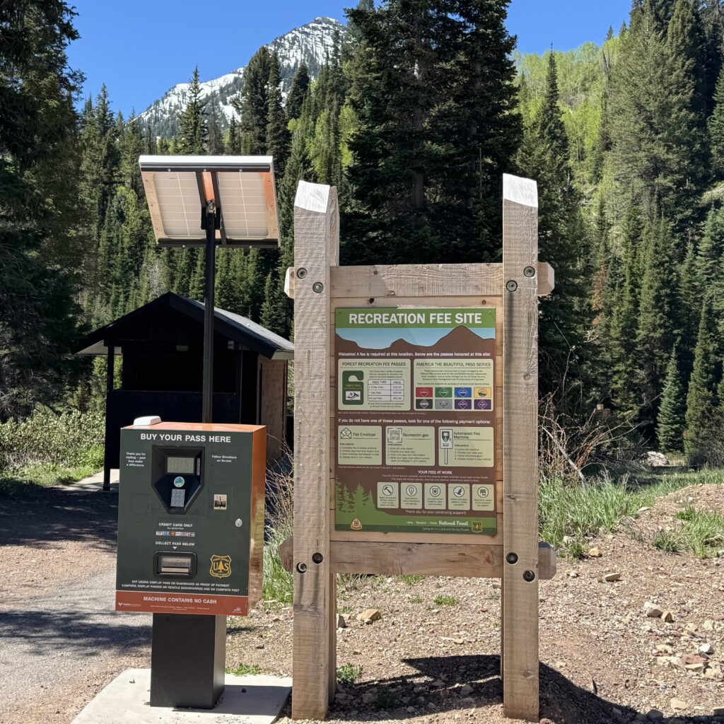 A U.S. Forest Service recreation fee station at Cardiff Fork stands beside a mountain trail in a forested canyon. The solar-powered payment kiosk, labeled โBuy Your Pass Here,โ is positioned next to a large wooden-framed sign explaining recreation fees and accepted passes. In the background, a snow-covered mountain peak rises above dense evergreen and aspen trees under a clear blue sky, highlighting the alpine setting.