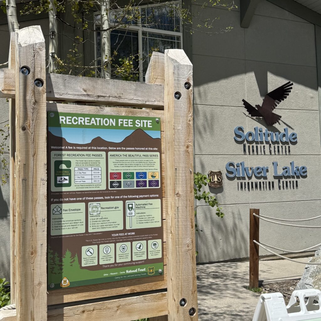 A U.S. Forest Service recreation fee information sign is displayed in a large wooden frame outside the Solitude Silver Lake Information Center. The sign outlines recreation fee requirements, accepted passes, and payment options for visitors. Behind it, the information center building features the โSolitude Silver Lake Information Centerโ name and bird logo, with aspens, landscaping, and a pedestrian walkway visible in the foreground.