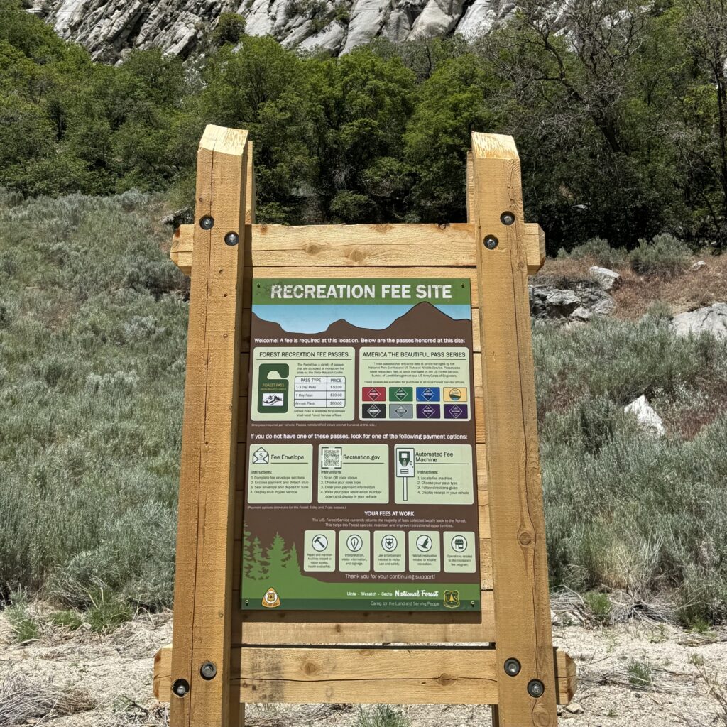 A U.S. Forest Service recreation fee information sign is mounted in a large wooden frame within a canyon landscape. The sign provides details about recreation fees, accepted passes, and payment options for visitors. Surrounded by sagebrush, rocky terrain, and dense green trees, the sign sits below a steep granite mountainside, highlighting the natural setting of the recreation area.