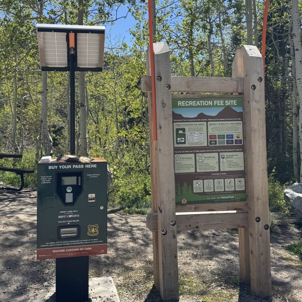 Alt-text: A solar-powered U.S. Forest Service recreation fee station at White Pine stands beside a wooden information sign in a forested mountain setting. The payment kiosk, labeled โBuy Your Pass Here,โ is mounted on a black post with solar panels above and accepts credit card payments for recreation fees. Next to it, a large wooden-framed sign displays information about recreation passes, accepted permits, and fee payment options. Aspen trees and green vegetation surround the site under a clear blue sky.