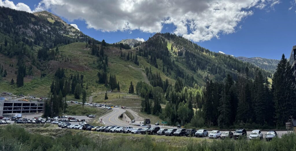 A line of parked cars along SR-210 in Little Cottonwood Canyon, with Snowbirdโs mountain slopes and a partly cloudy sky in the background.